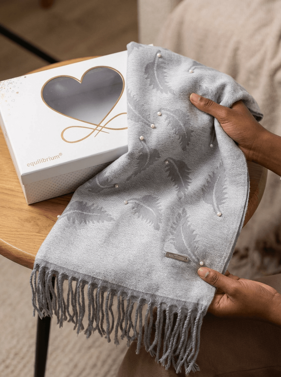 Gray scarf with fringe held by a hand, next to a white box with a heart design on a wooden surface.