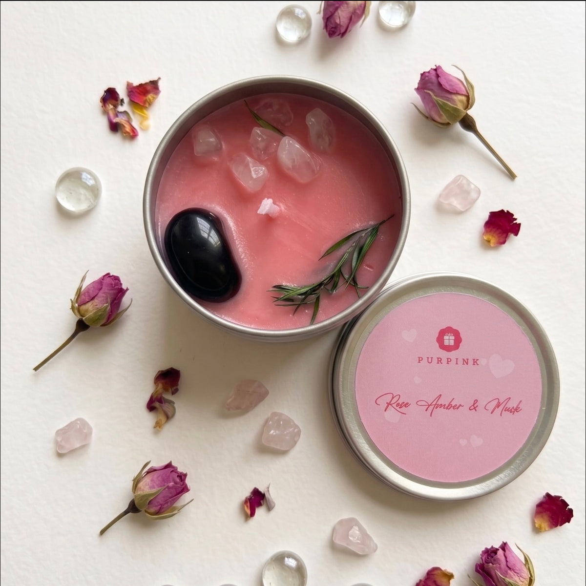 A pink wax candle in a round metal tin with its lid placed beside it on a white surface. The lid label reads PURPKINK and Rose Amber & Mint.