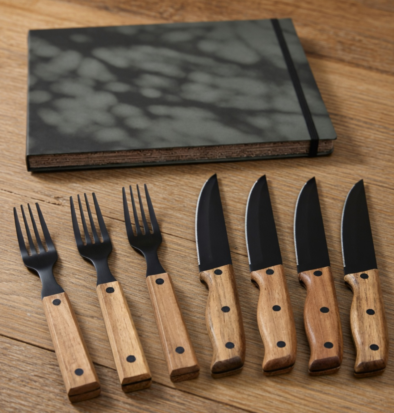 Set of knives and forks with wooden handles on a wooden surface, next to a black notebook.