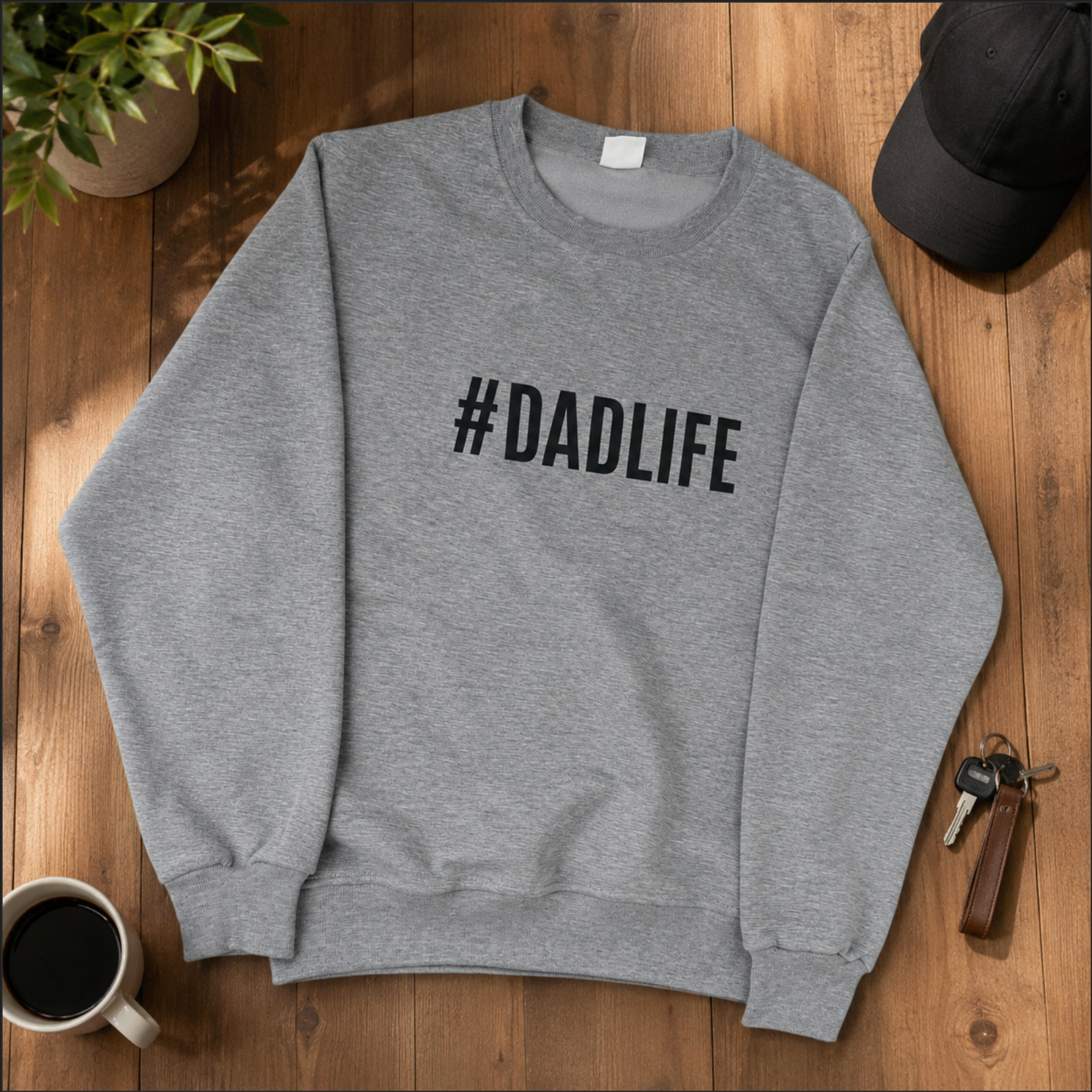 Gray sweatshirt with '#DADLIFE' text on a wooden surface with a cup of coffee, keys, and a cap.