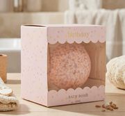 Bath bomb in a 'Happy Birthday' themed packaging on a wooden surface.