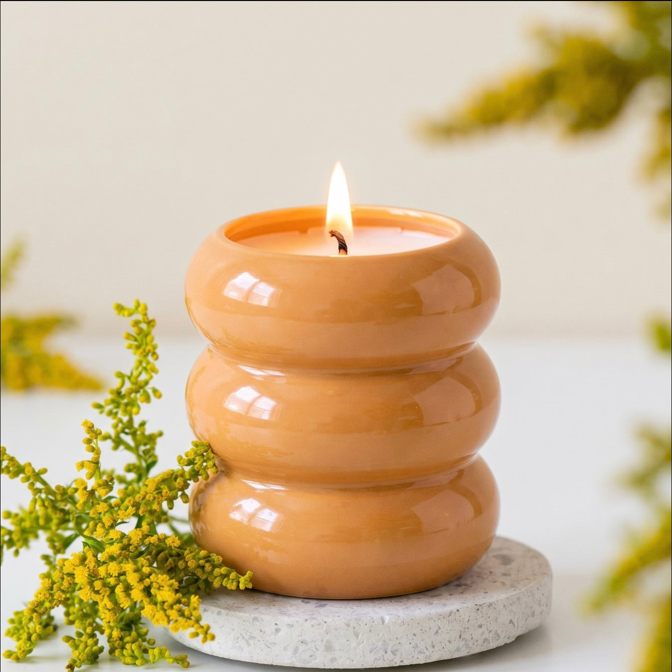 A three-tiered glossy ceramic candle holder in a warm terracotta orange color, lit with a small flame and styled with yellow flowers on a light gray stone coaster against a soft beige background.