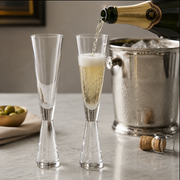 Two clear champagne flutes on a marble countertop, with one filled with sparkling champagne and the other empty. A bottle of champagne is pouring into the flute, with a metal ice bucket, cork, and olives in the background.
