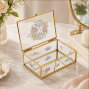 A rectangular glass jewelry box with a gold metal frame, shown open on a light marble surface. The clear glass panels feature delicate floral and botanical prints in soft pastel tones, and the interior has three compartments.