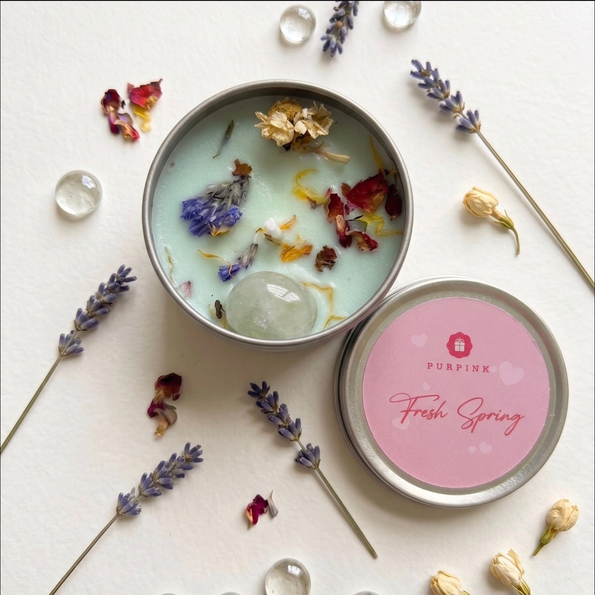 A round metal tin of scented candle with its lid beside it, styled on a white surface with dried lavender sprigs, flower petals, and clear glass beads. The candle wax is pale mint green and contains dried botanicals and a clear stone.