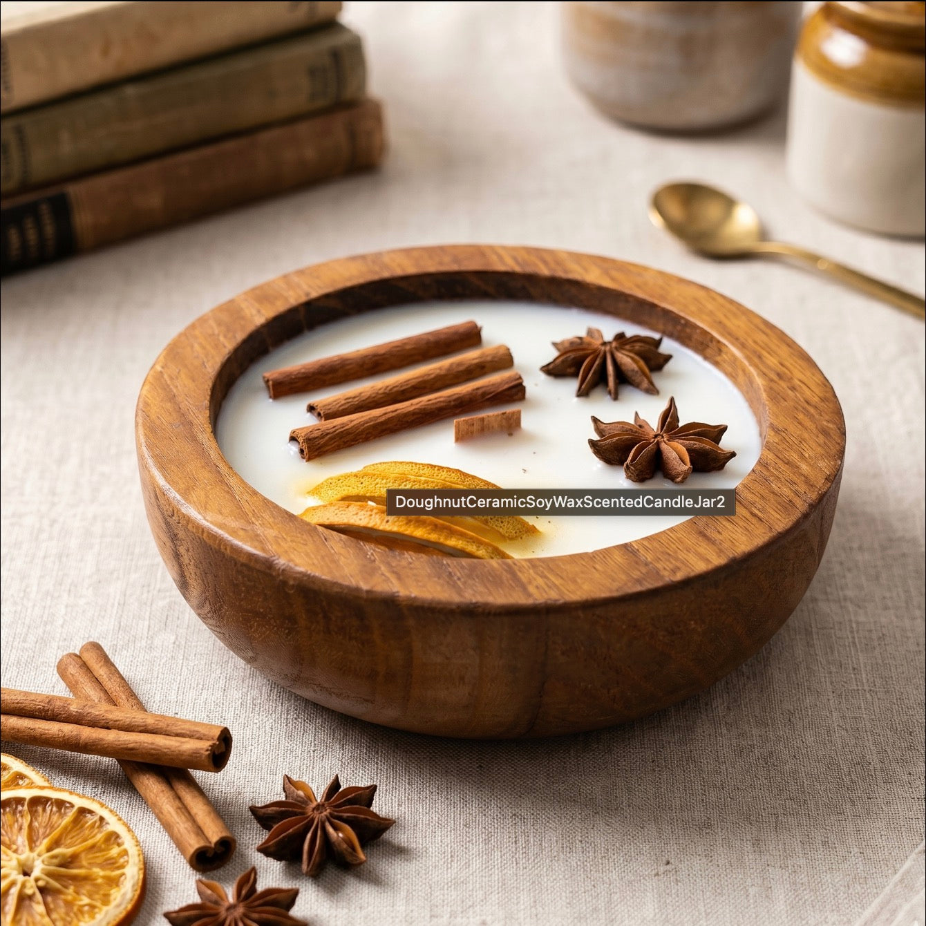 A round wooden bowl filled with a white scented candle, decorated with cinnamon sticks, star anise, and dried orange slices, set on a linen surface with books and a spoon in the background.