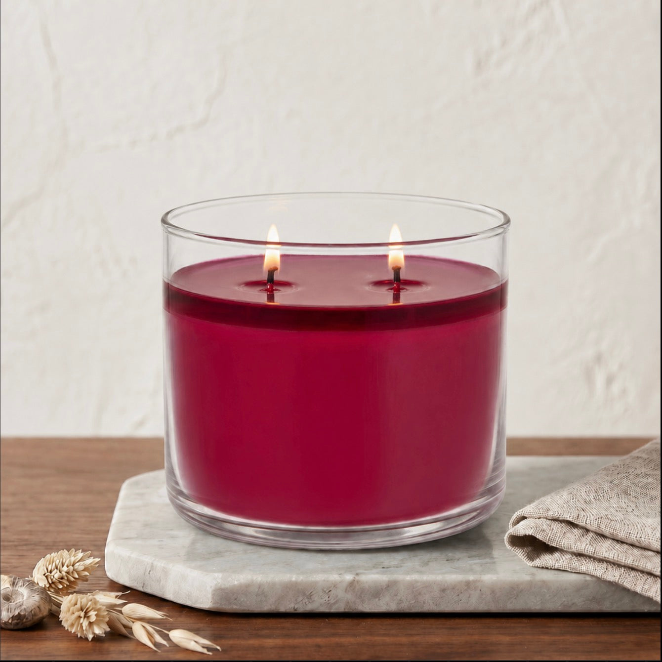 A double-wick candle in a clear glass jar with deep red wax, lit and set on a gray marble tray with dried wheat and a folded linen cloth against a textured off-white wall.