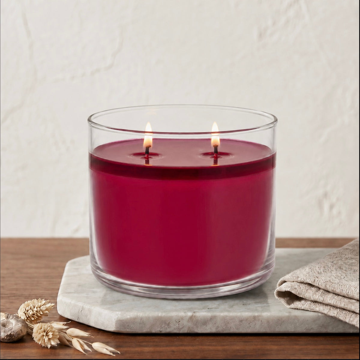 A double-wick candle in a clear glass jar with deep red wax, lit and set on a gray marble tray with dried wheat and a folded linen cloth against a textured off-white wall.