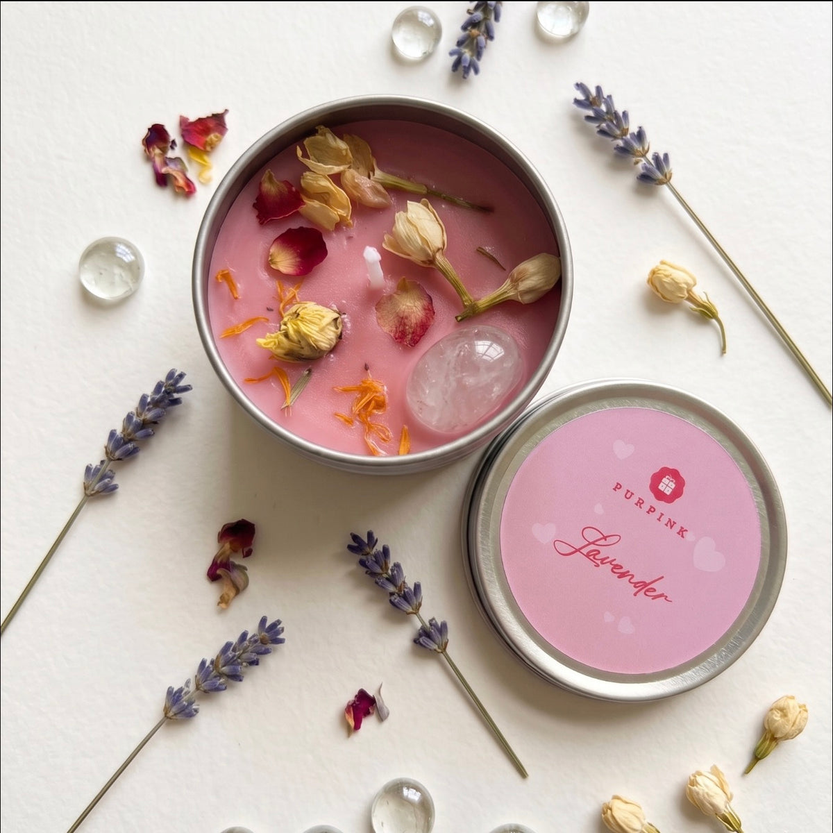 An open round metal tin candle with pink wax sits on a white surface, surrounded by lavender sprigs, dried flower petals, and clear glass beads. The lid rests beside it, showing a pink label that reads PURPLINK and Lavender.