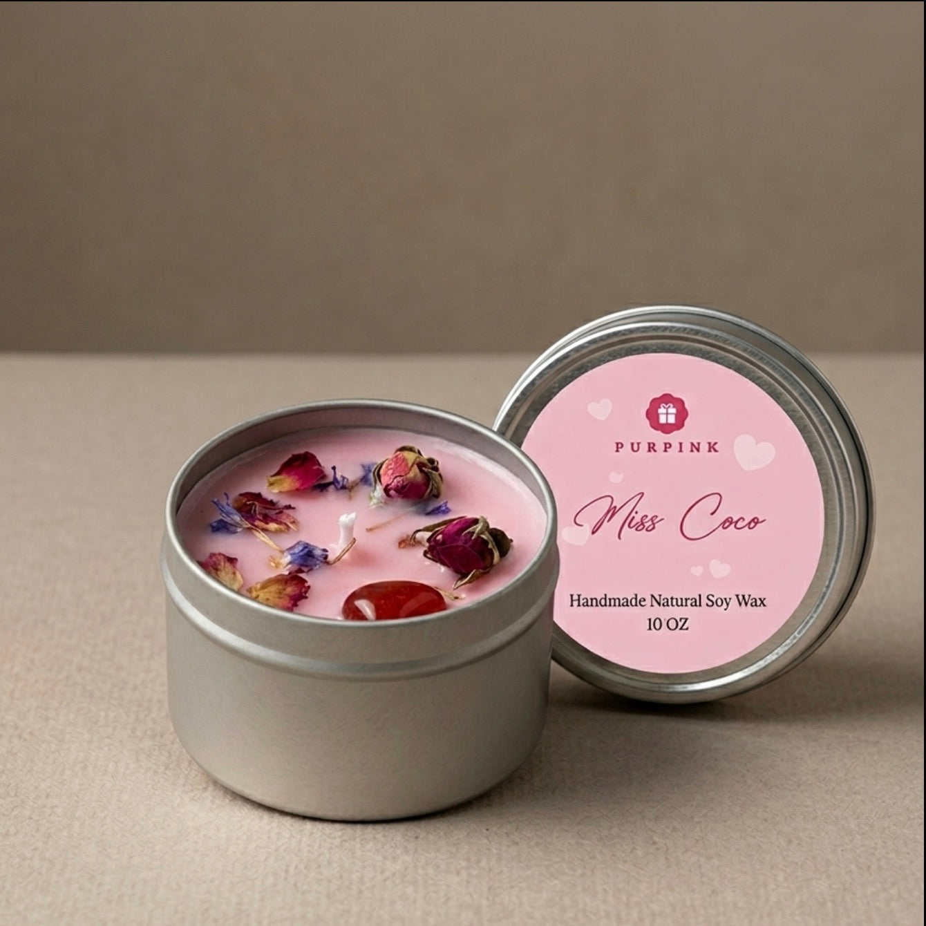 An open round silver tin of PURPINK Miss Coco handmade natural soy wax candle, showing pink wax with dried rose petals, next to its pink lid on a neutral beige background.