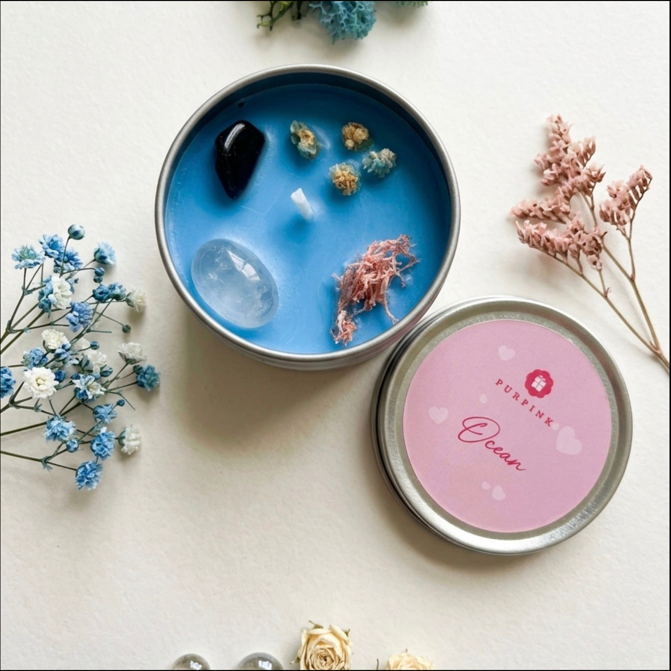 A round metal candle tin shown open with blue wax inside, surrounded by dried flowers and crystals. The lid sits beside it on a light beige surface, featuring a pink label with the brand name PURFINK and the scent name Ocean.