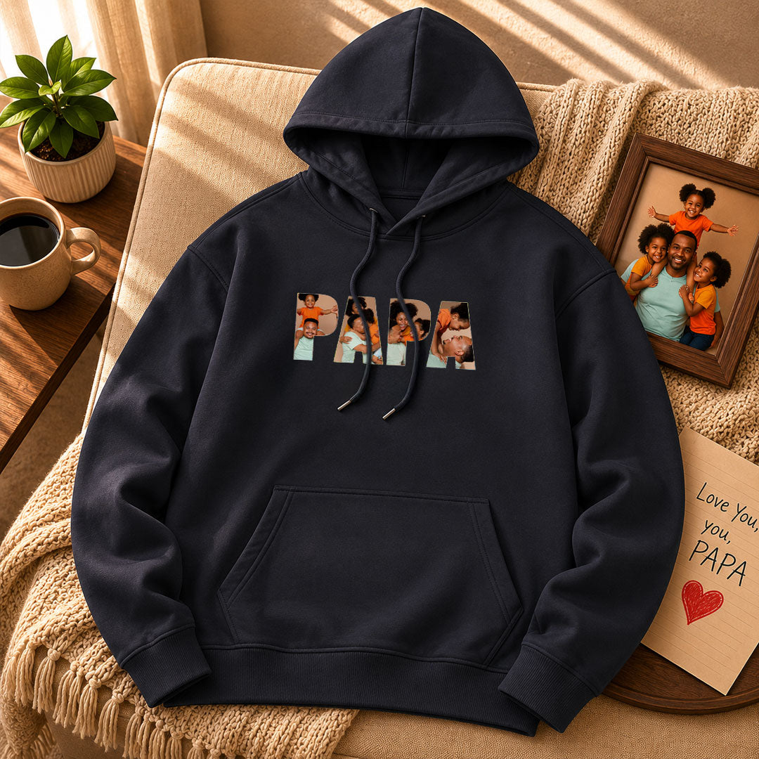 Black hoodie with 'PAPA' graphic on a beige couch with a cup and photo frame in the background.