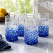 Four clear glass tumblers with blue speckled patterns near the bottom, shown on a marble countertop with water being poured into them from a clear pitcher. A blurred kitchen background with lemons is visible.