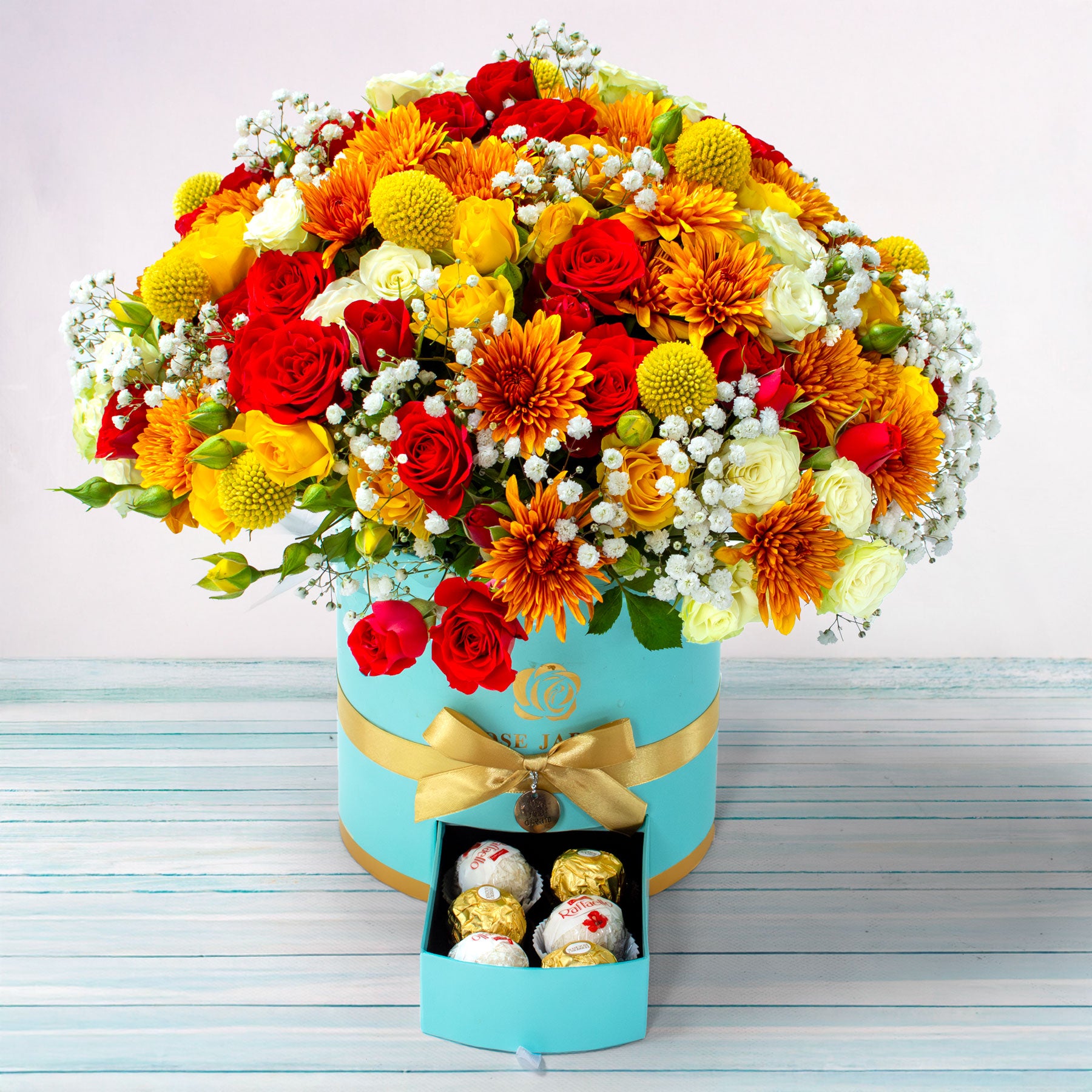 Colorful bouquet of flowers in a turquoise box with gold ribbon, accompanied by a small box of chocolates on a light wooden surface.