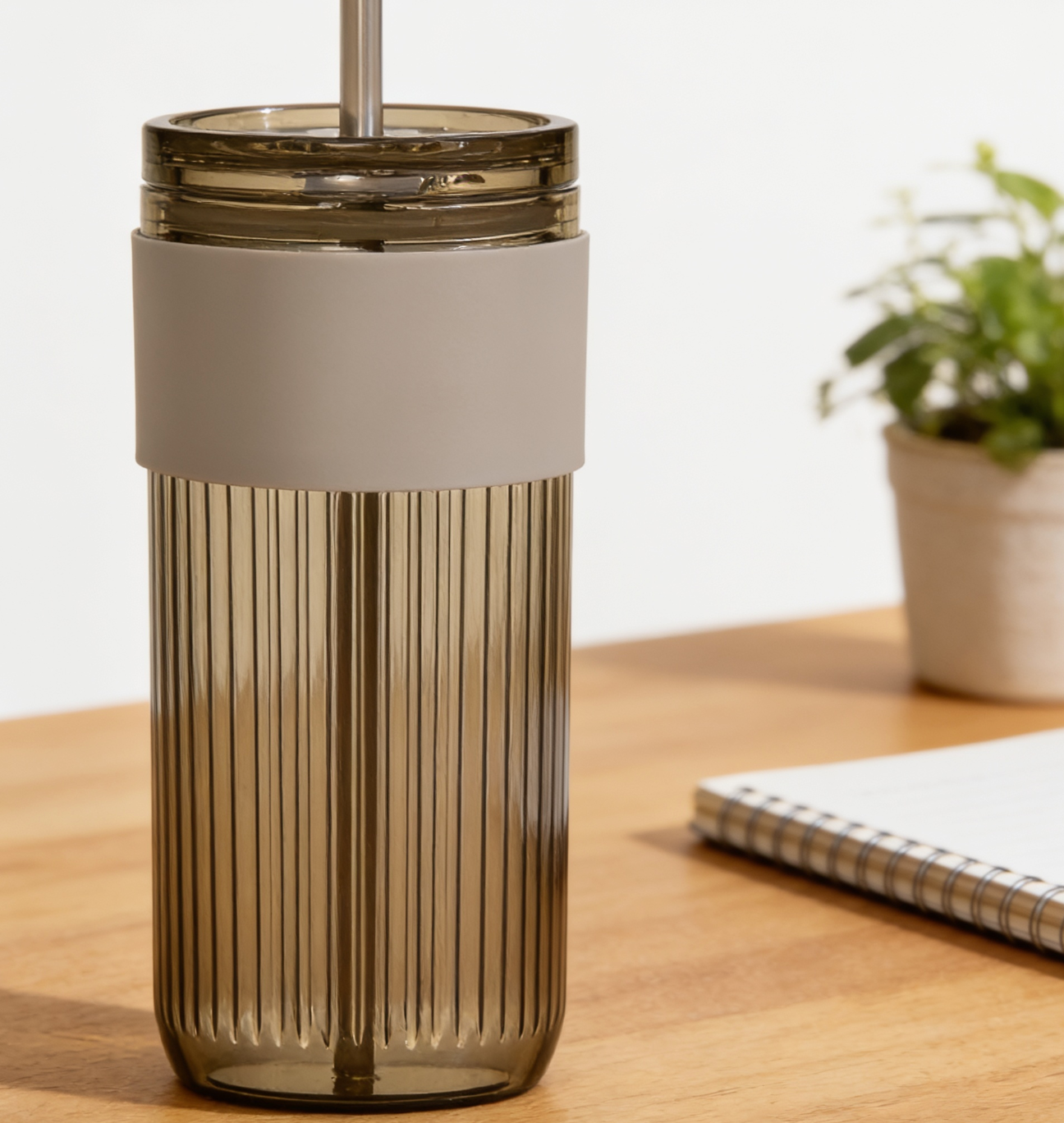 Clear tumbler with beige lid on a wooden surface with a plant and notebook in the background