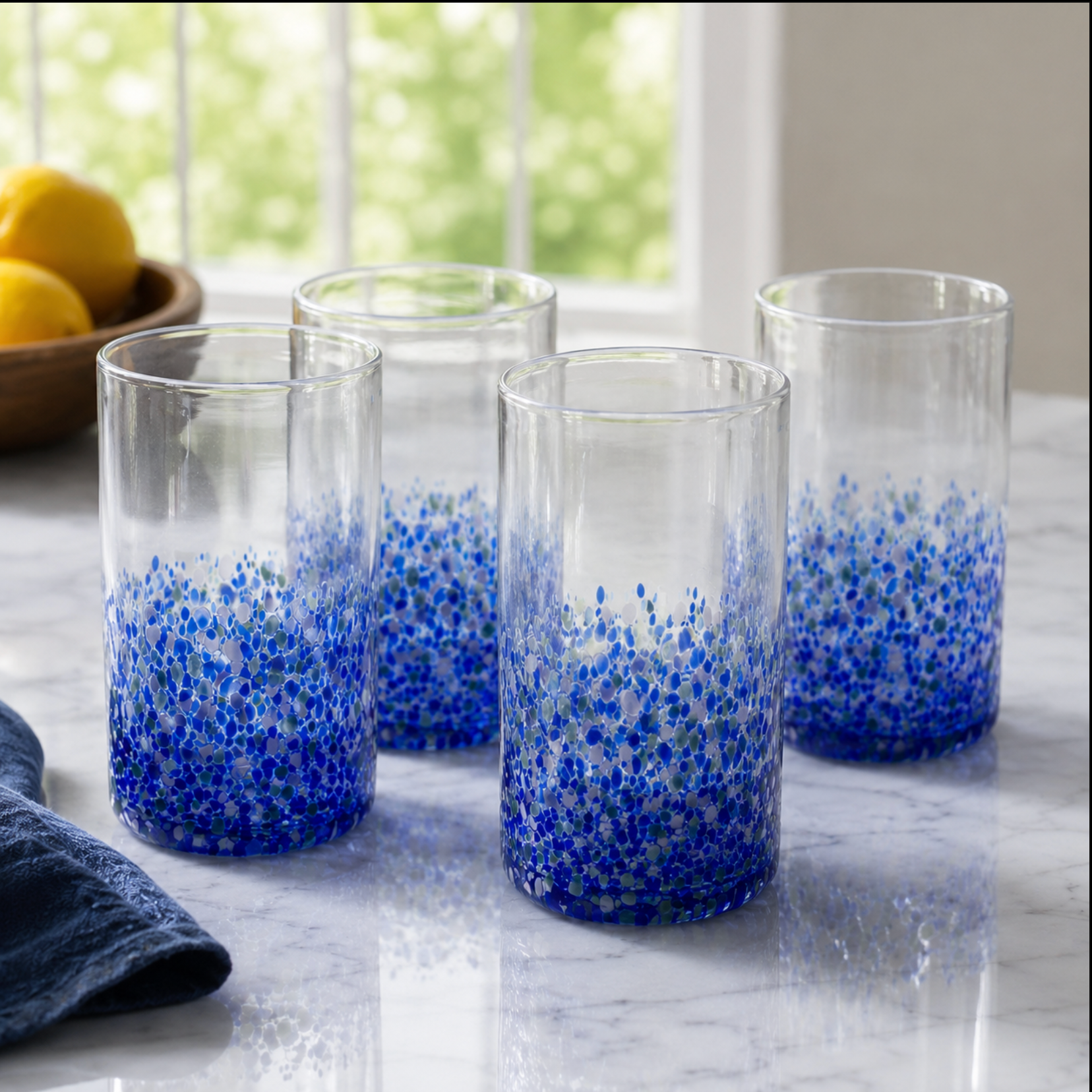 Four clear glass tumblers with a dense blue speckled pattern concentrated at the bottom, arranged on a light marble countertop near a window.