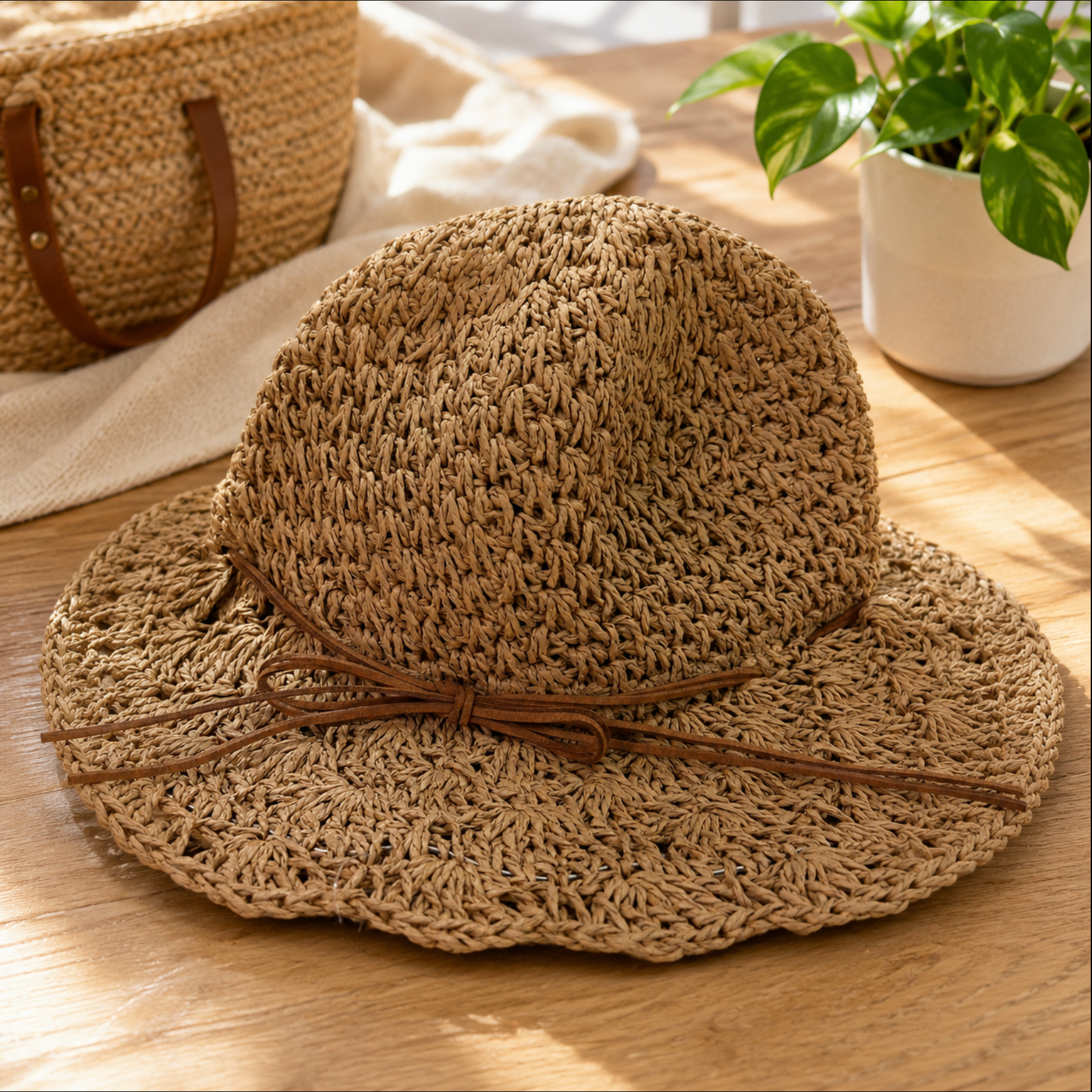 A natural woven straw sun hat with a wide brim and a brown ribbon bow, displayed on a light wooden table with a potted plant and a woven bag in the background.