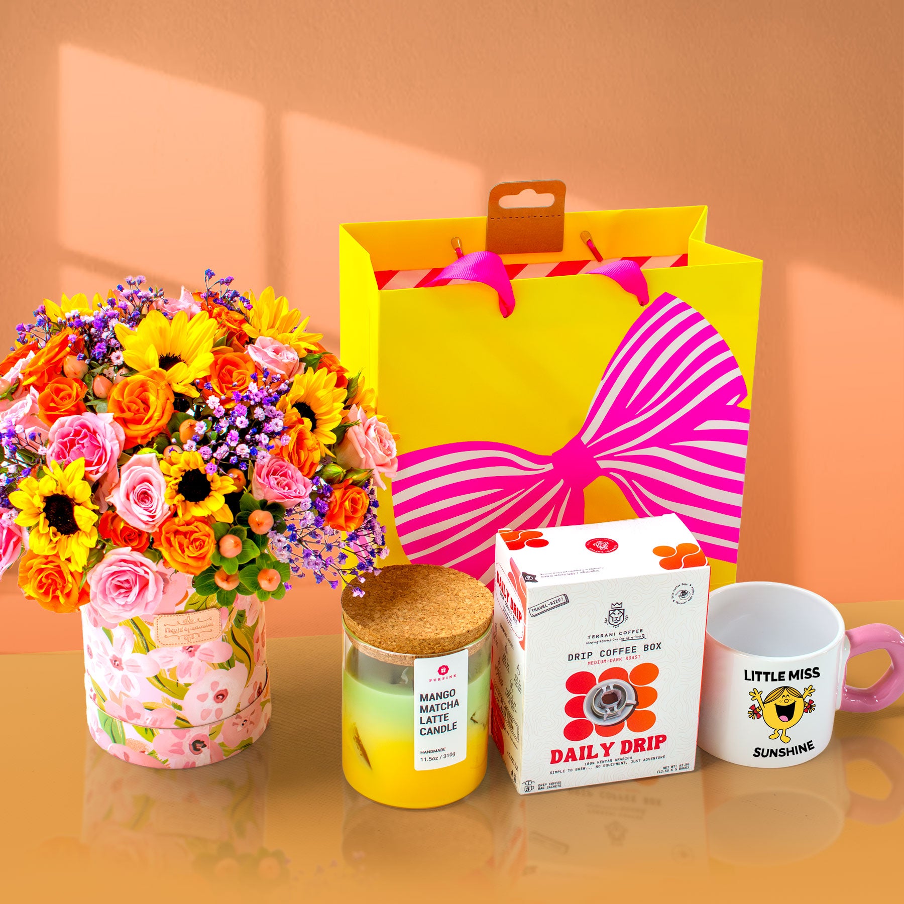 Floral arrangement, yellow candle, coffee box, and mug on a reflective surface with a pink background