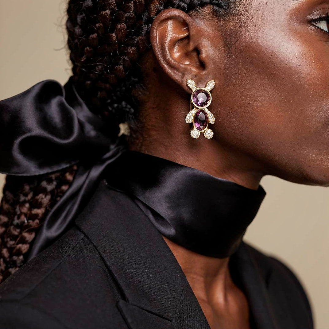 Close-up of a person wearing a decorative earring with a purple gemstone on a neutral background