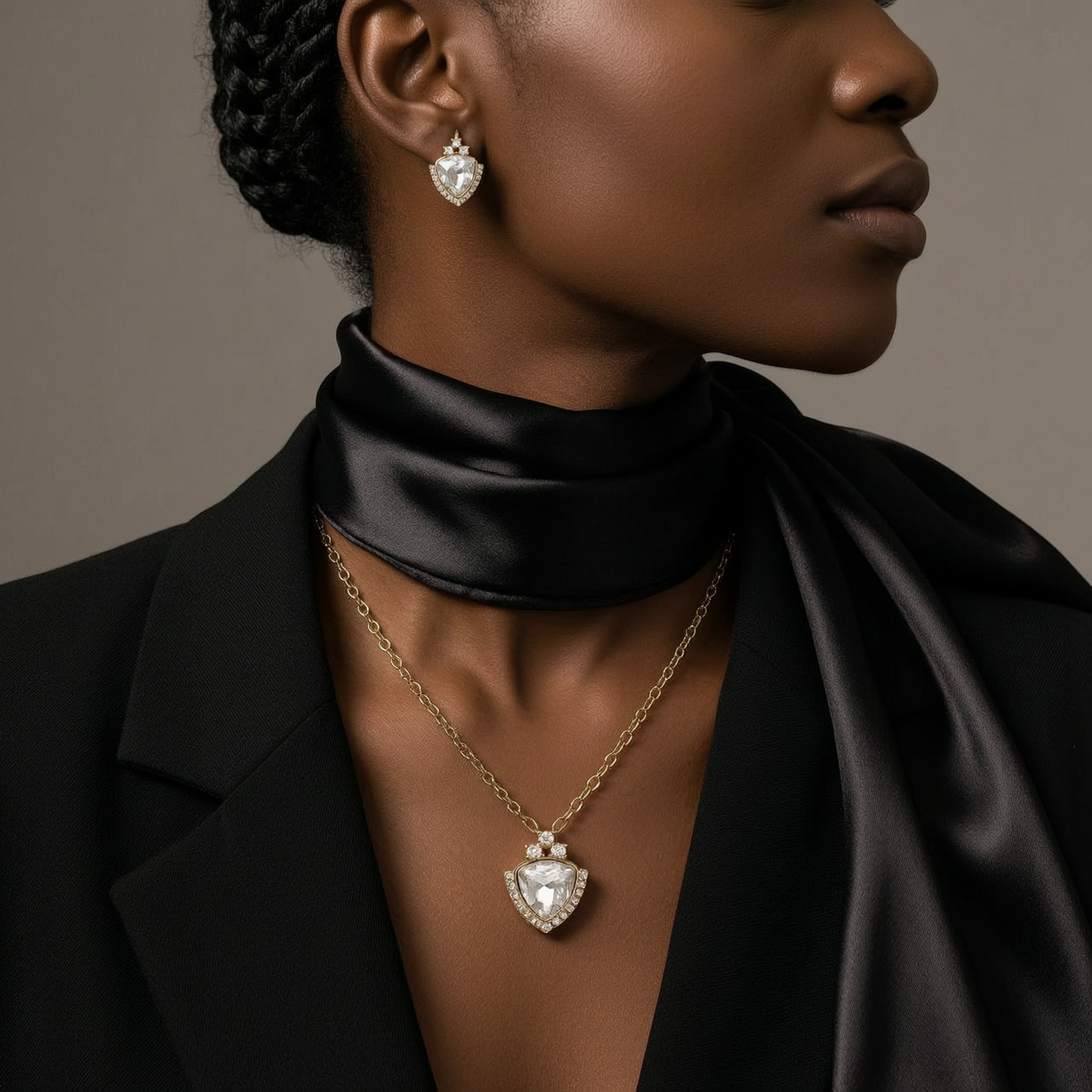 Woman wearing a black garment, gold necklace with a heart-shaped pendant, and diamond earrings against a neutral background.
