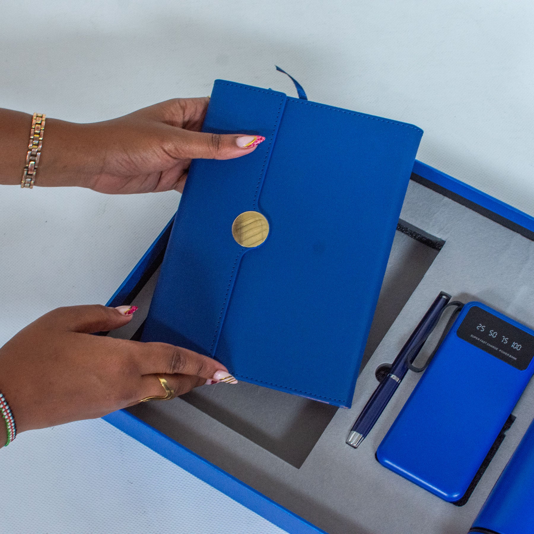 A blue notebook with a gold button is held by a hand, with a phone and pen on a grey surface.