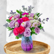 Colorful flower bouquet in a gradient purple and blue vase on a woven tray with a white background