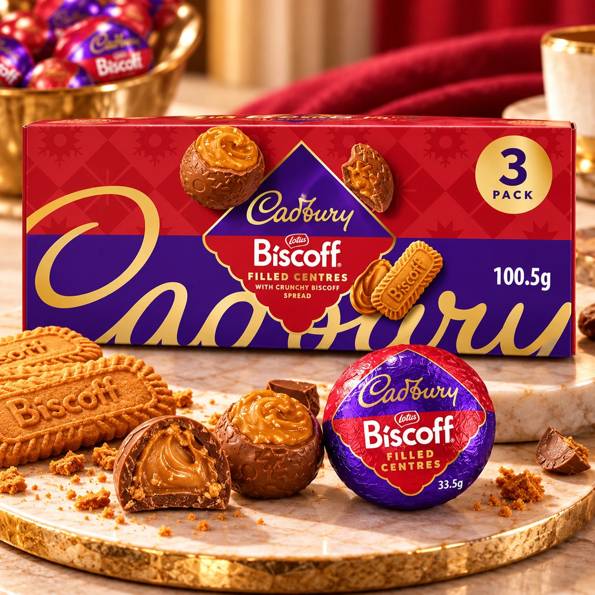 A Cadbury Biscuff filled centres 3-pack box of spread, shown with several wrapped and unwrapped chocolates and biscuit pieces on a marble board in a styled setting.