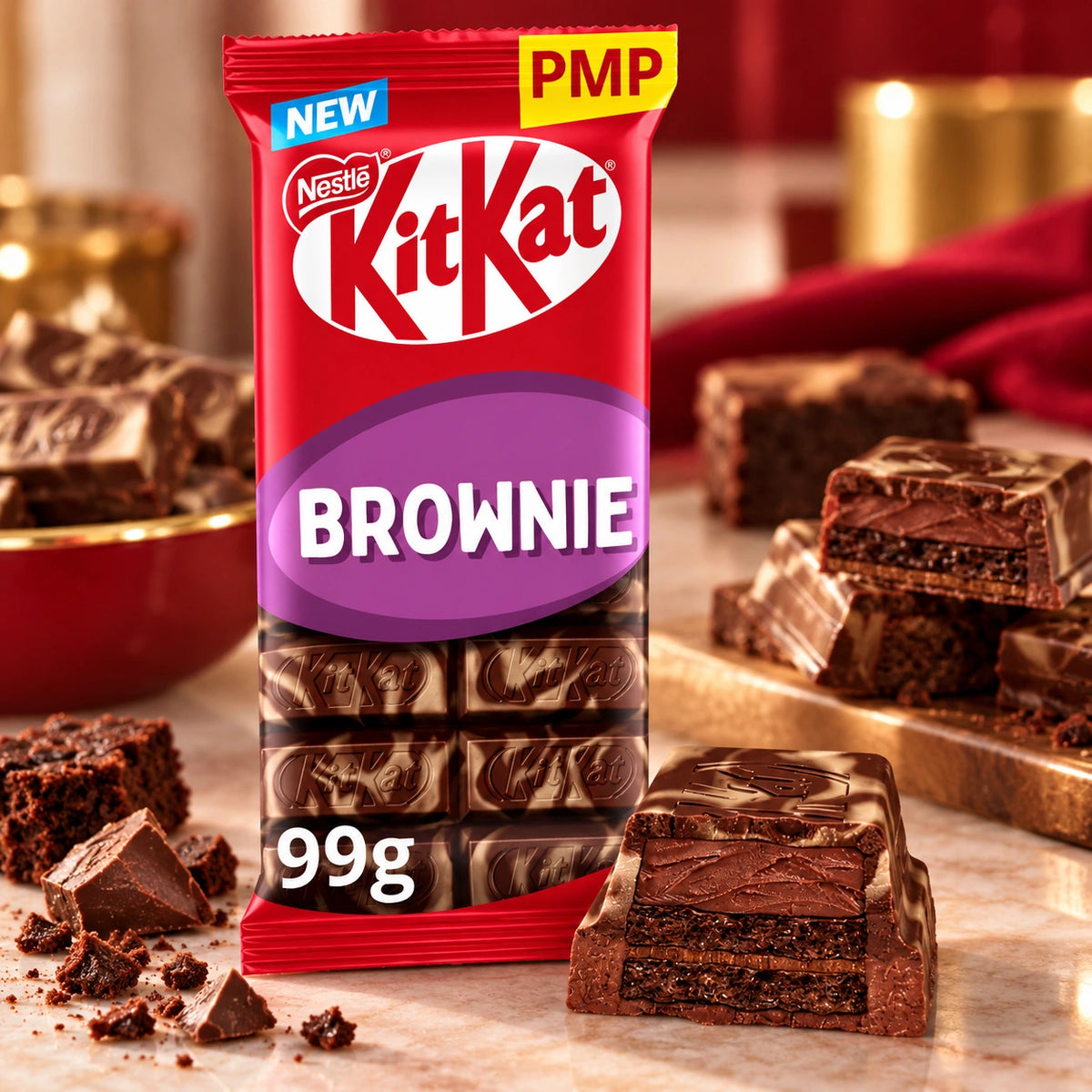 A package of KitKat Brownie chocolate bars stands on a marble surface with brownie pieces arranged around it. The front of the bag shows the KitKat logo, a blue NEW banner, a yellow PMP sticker, and the word BROWNIE.