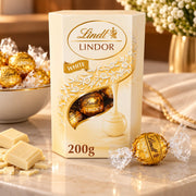 A 200g box of Lindt Lindor White chocolate, shown on a marble surface with white chocolate pieces and wrapped chocolates in the background.